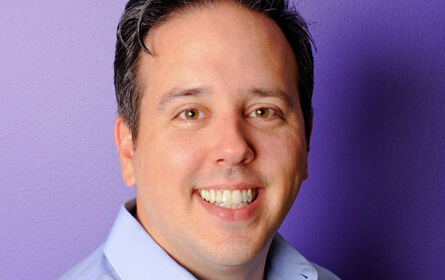 A man smiling in front of a purple wall.
