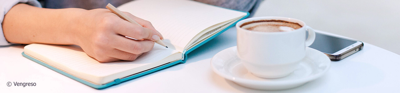 close up of a hand writing on an open notebook on a desk next to a cup of coffee