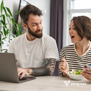 a man with a beard and tattoos sitting in front of an open laptop looking at the woman sitting next to him who is screaming and smiling with her hands up on the table