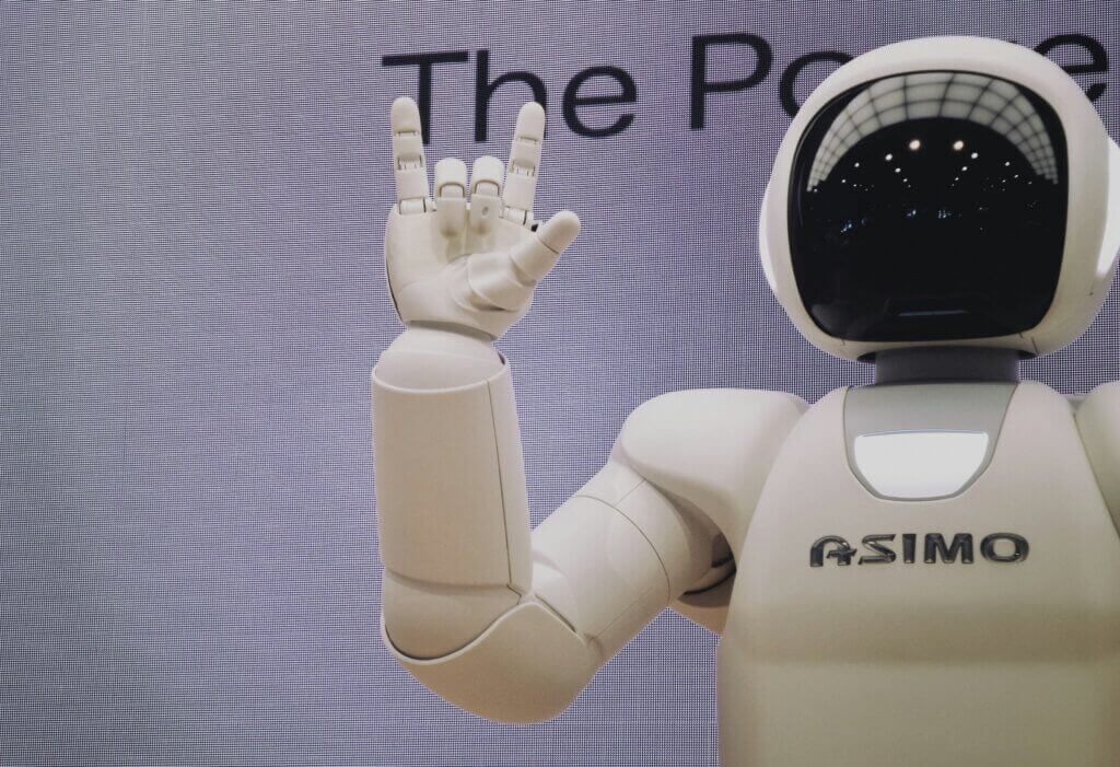 A humanoid robot with a white exterior and a glossy black faceplate stands and raises its left hand, making the "rock on" gesture. The AI assistant has "ASIMO" printed on its chest. The background is a light gray screen displaying partial text "The Pow".