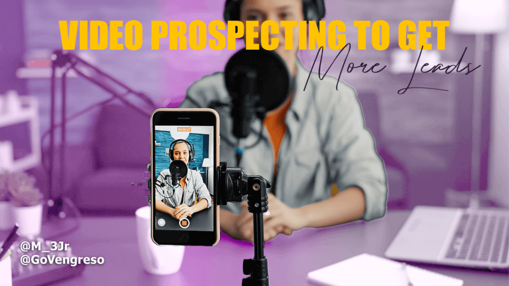 A person in a casual shirt sits at a table with a microphone, engaging in video prospecting toward a smartphone on a tripod. The phone displays their image, with text at the top reading "VIDEO PROSPECTING TO GET MORE LEADS." Social media handles and a coffee cup adorn the scene.