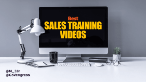 A desktop setup features a large monitor showing "Best SALES TRAINING VIDEOS" in bold yellow and red. Nearby, a desk lamp, notebook, black mug, tablet, mouse, keyboard, and small plant complete the scene. The handles @M_3Jr and @GoVengreso rest at the bottom left against a gray background.